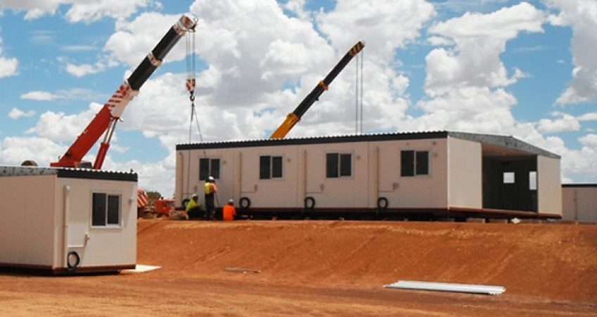 Modular commercial buildings being assembled on site with the help of cranes under a clear sky.