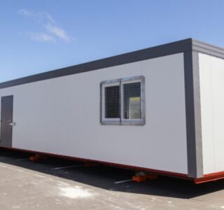 Portable gray office container with windows and doors, set on orange skates in a parking area.