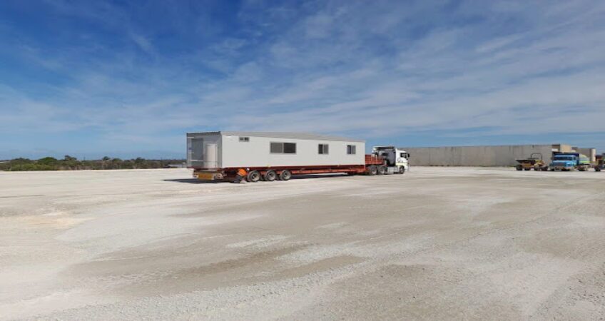 Truck transporting a portable modular office building on a flatbed trailer across a large, open area.