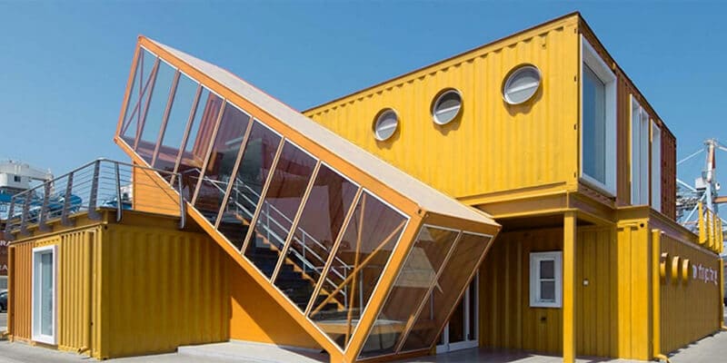 Yellow shipping container conversions with circular windows and an angled glass staircase structure.